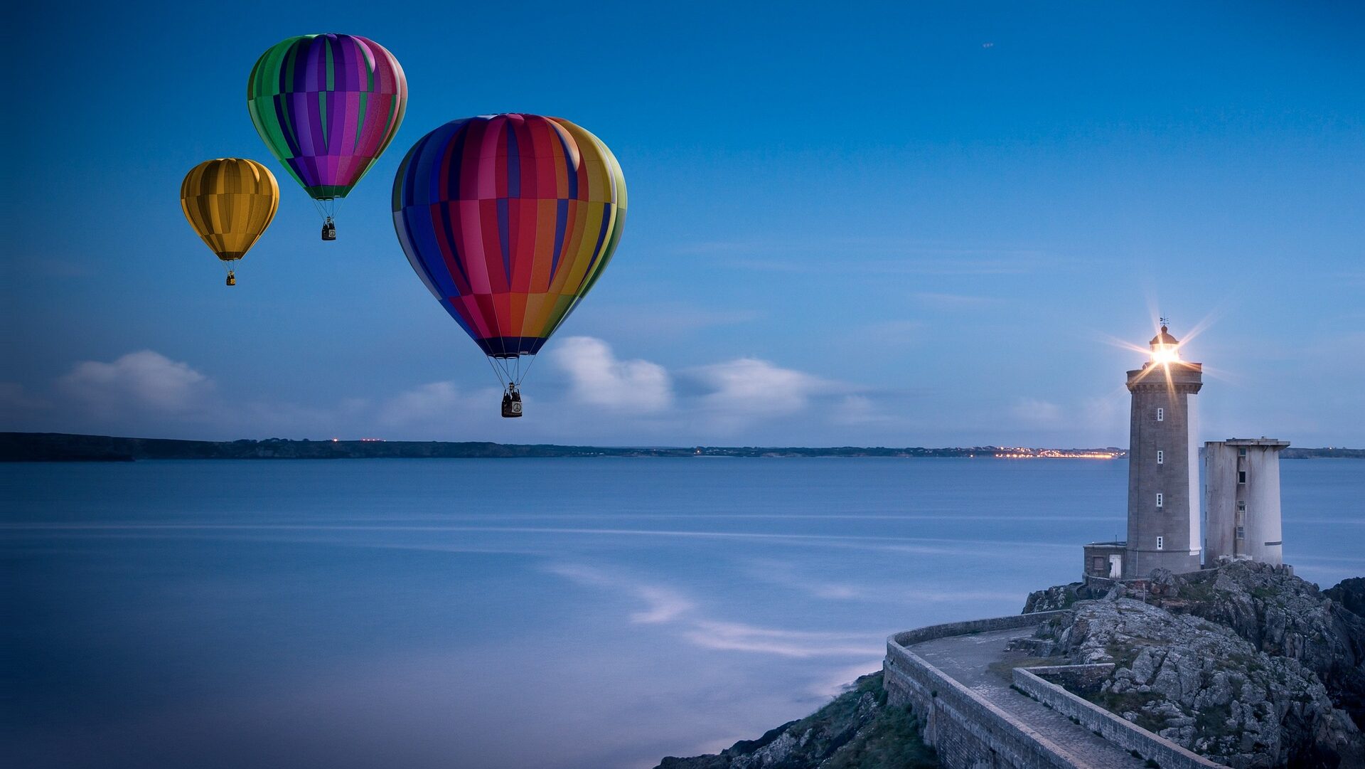 Three hot air balloons over water approaching a lighthouse on a rocky sea cliff.