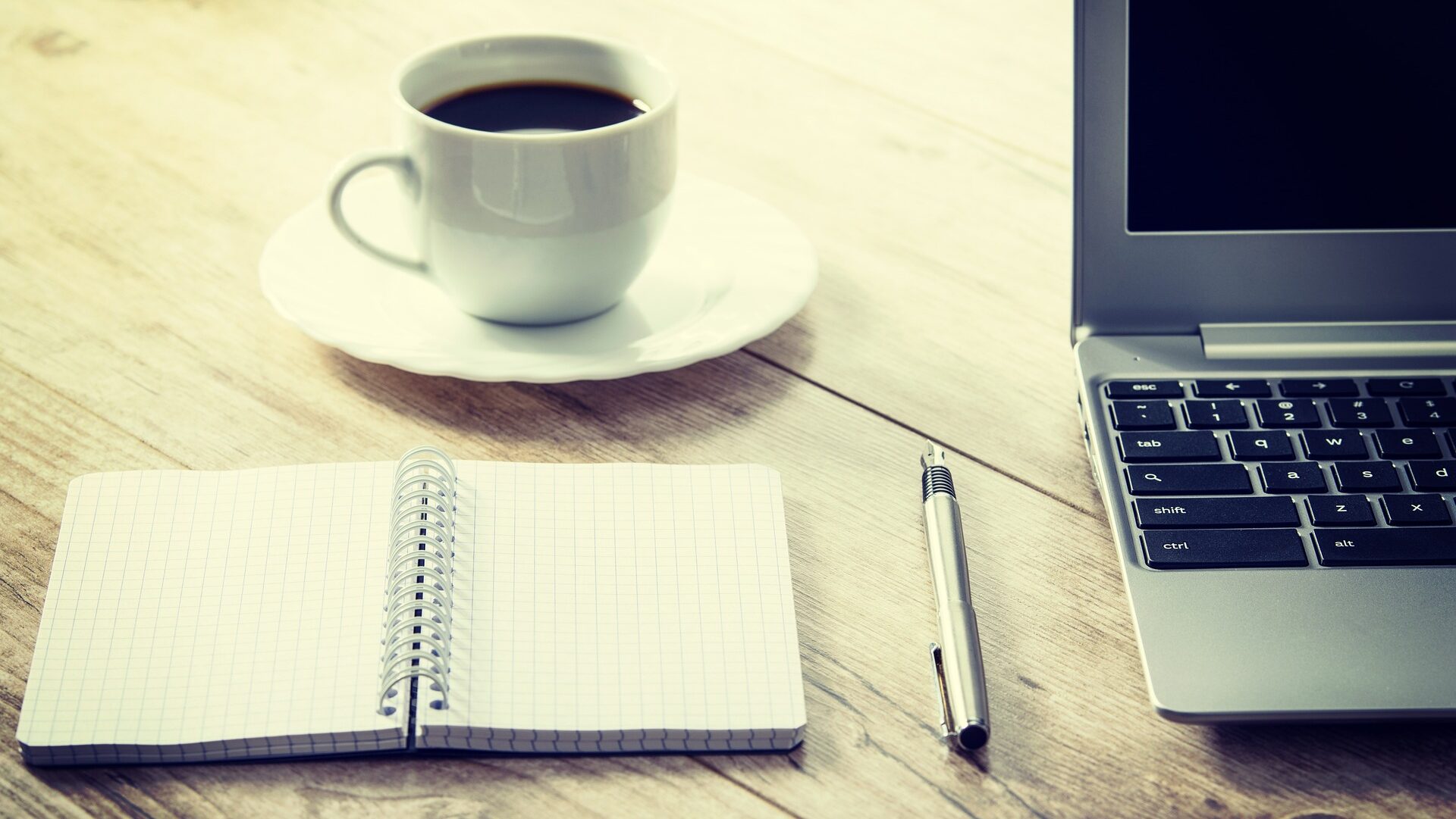 Coffee in a white mug on a saucer, with a notebook, pen, and laptop on a table