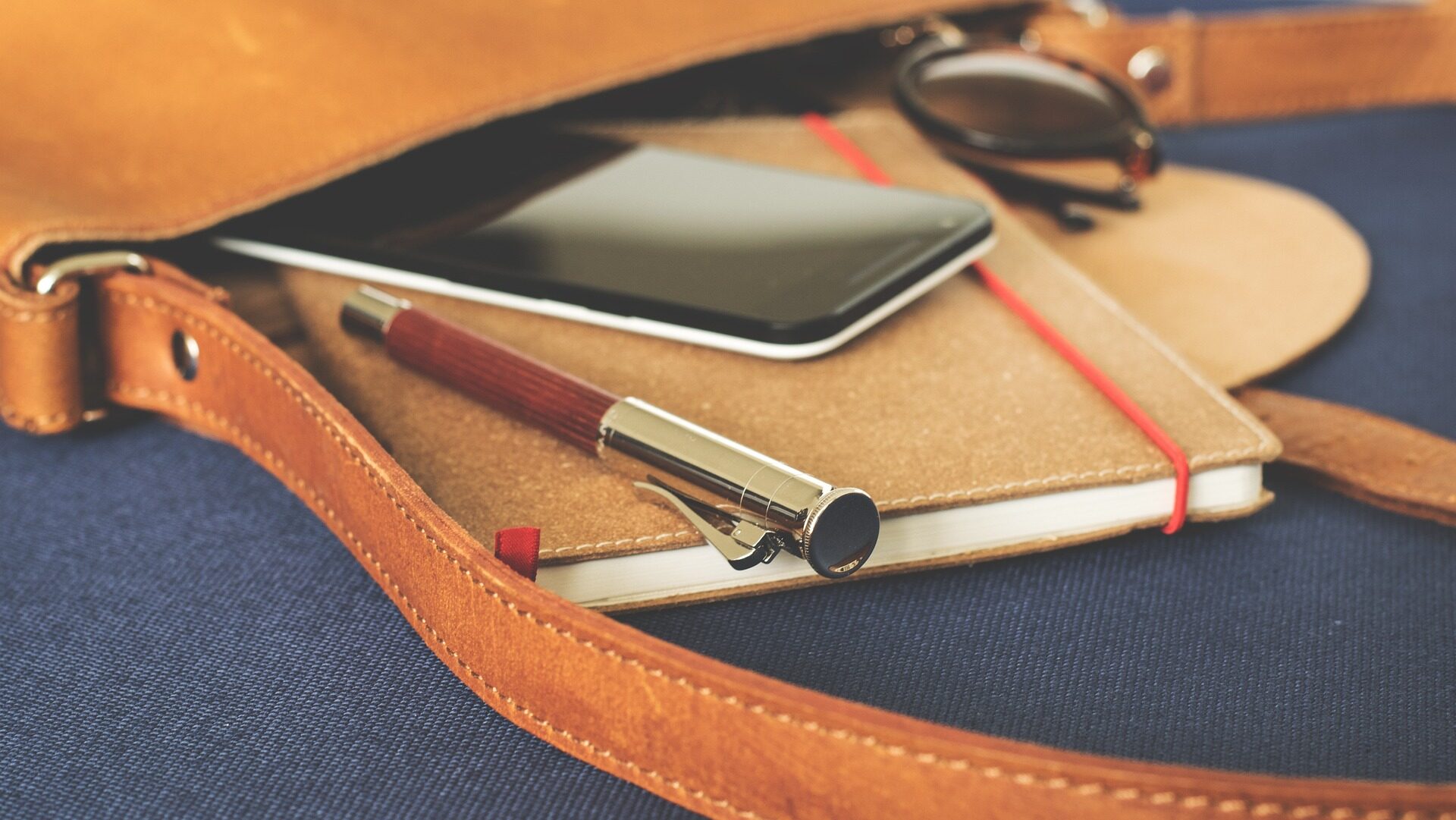 Leather bag with notebook, pen, phone, and sun shades.